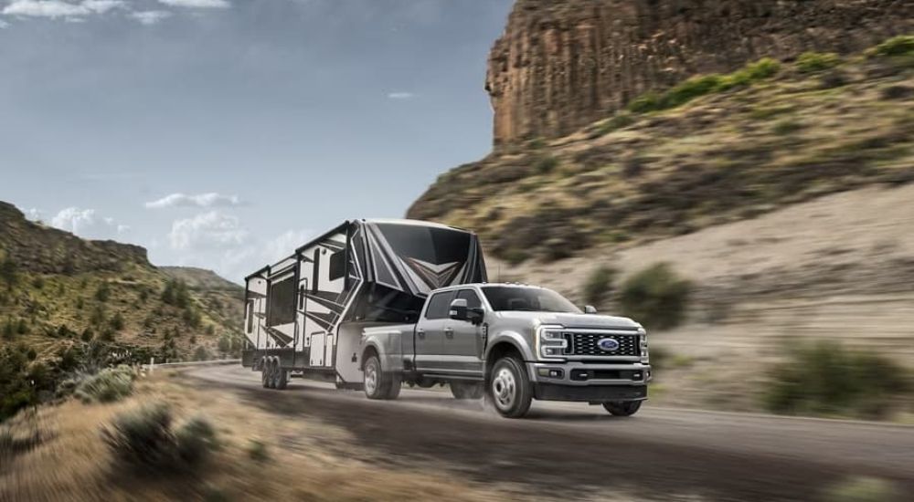 A silver 2023 Ford Super Duty tows a camper leaving a Ford truck dealer. 