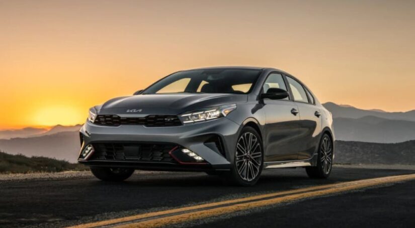 A silver 2023 Kia Forte for sale is shown driving along a highway at sunset.