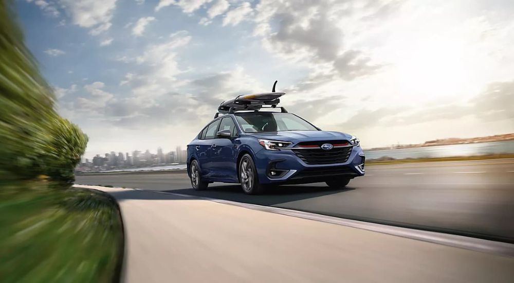 A blue 2023 Subaru Legacy with a surfboard in the roof rack is shown driving next to a river. 