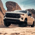 A white 2023 Chevy Silverado 1500 Z71 Custom Trail Boss is shown climbing over rocks in the desert after leaving a Chevy Silverado dealer.