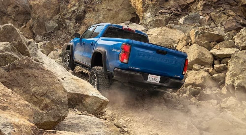A blue 2023 Chevy Colorado is shown from behind driving up a rocky and dusty trail. 