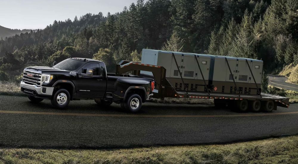 A black 2023 GMC Sierra HD is shown towing a flatbed trailer with large units on it up a winding mountain road. 