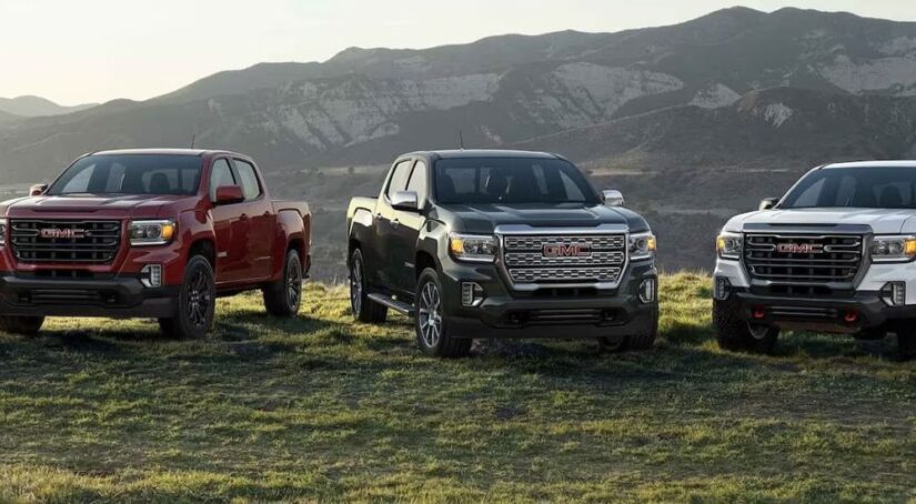 Three GMC trucks for sale in red, black, and white are parked on a hilltop in front of a mountain range.