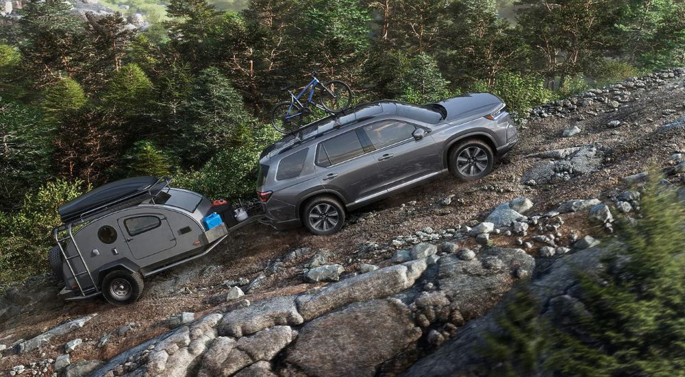 A gray 2023 Honda Pilot towing a matching small camper is shown driving up an off-road trail.