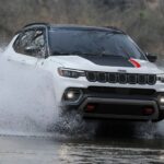 A white 2023 Jeep Compass is shown driving through a river sending water spraying into the air on either side.