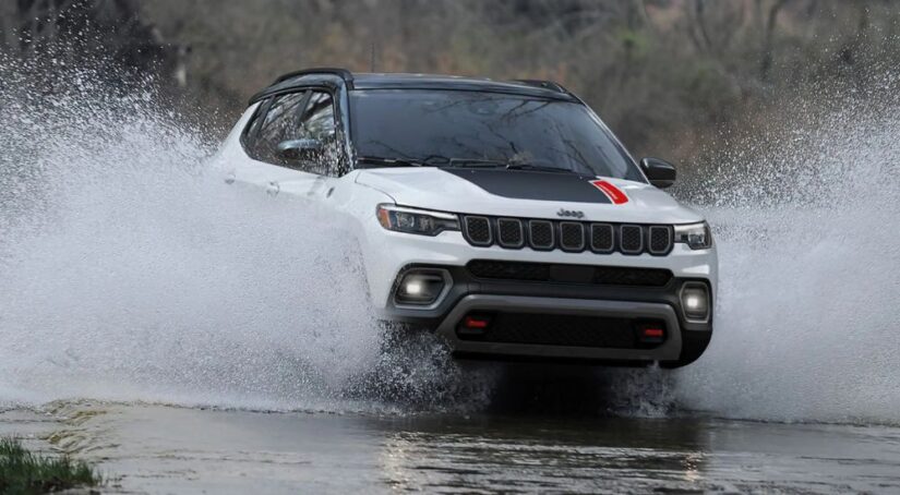 A white 2023 Jeep Compass is shown driving through a river sending water spraying into the air on either side.