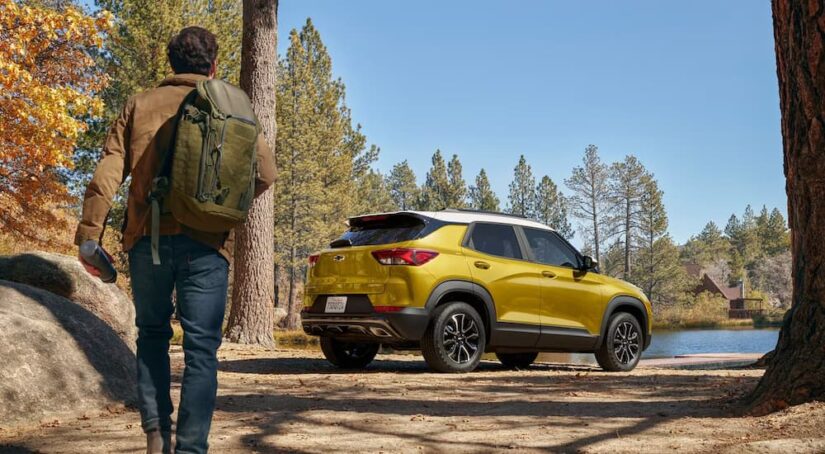 A man with a backpack is shown approaching a yellow 2023 Chevy Trailblazer parked by a lake in the woods.