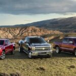 A red 2015 Chevy Colorado, black 2015 Chevy Silverado HD, and red 2015 Chevy Silverado 1500 are shown parked side by side in an open field with mountains in the background.