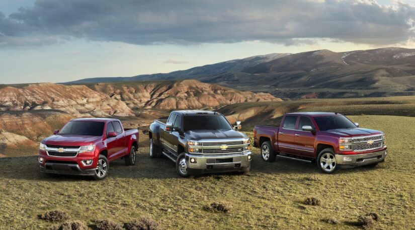A red 2015 Chevy Colorado, black 2015 Chevy Silverado HD, and red 2015 Chevy Silverado 1500 are shown parked side by side in an open field with mountains in the background.