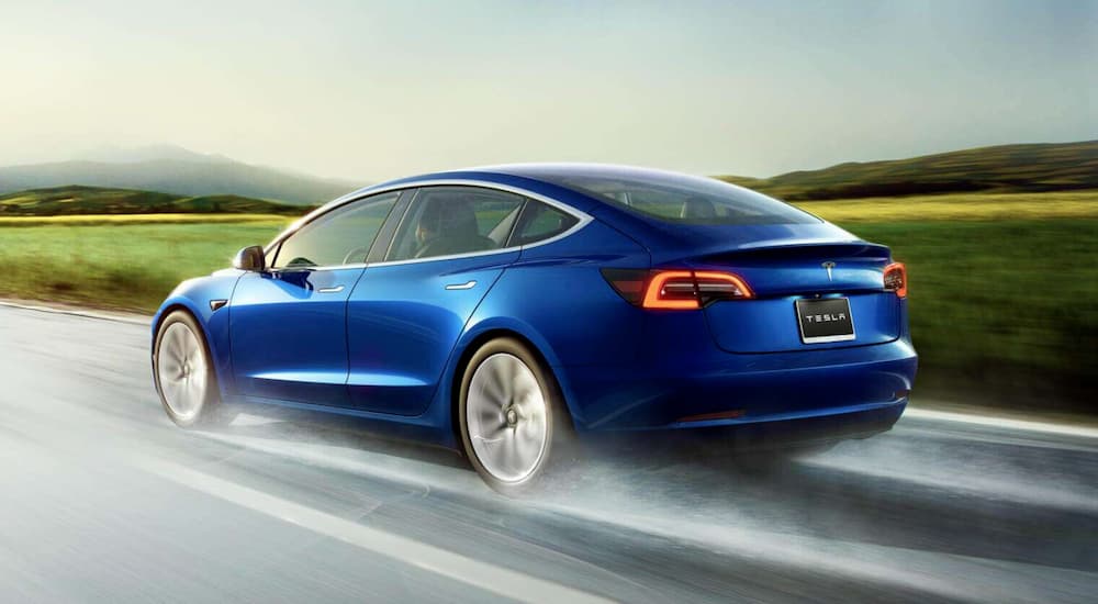 A blue 2020 Tesla Model 3 is shown driving on a wet road.