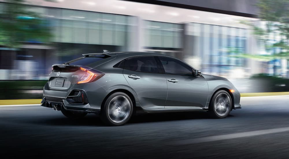 A silver 2021 Honda Civic Hatchback Sport Touring is shown driving on a road near a building.