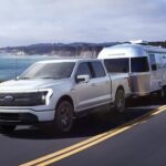A white 2023 Ford F-150 Lightning is shown driving on a highway towing a trailer.
