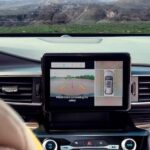 The black interior of a 2023 Ford Explorer XLT is shown from the backseat. A woman in the drivers seat watches a rearview camera on the dashboard.