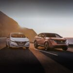 Two popular electric vehicles for sale, a white 2023 Nissan LEAF and an orange 2023 Nissan Ariya, are shown driving beside each other on an oceanside road.