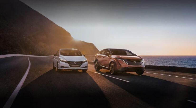 Two popular electric vehicles for sale, a white 2023 Nissan LEAF and an orange 2023 Nissan Ariya, are shown driving beside each other on an oceanside road.