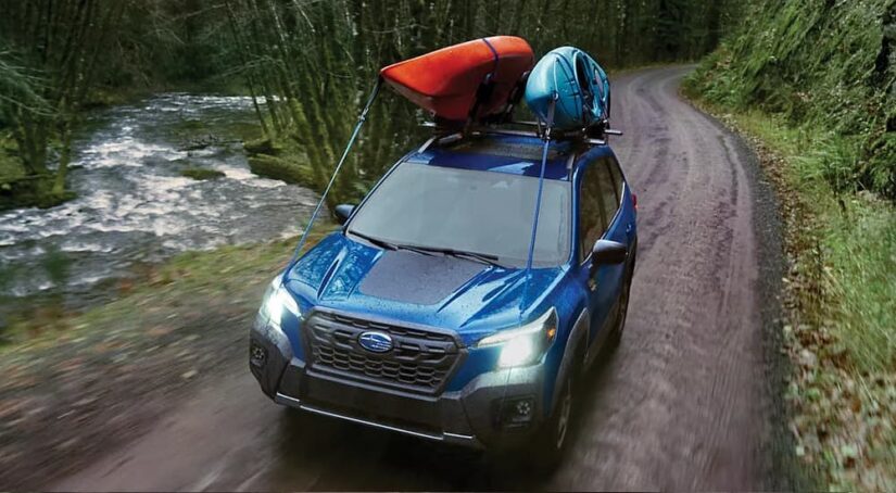A new Subaru Forester for sale, a blue 2023 Subaru Forester Wilderness, is shown driving through a forest with kayaks strapped to the top of it.