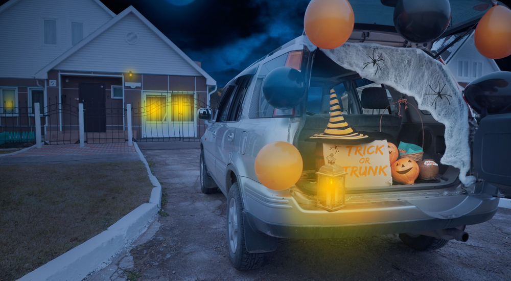 A white SUV with an open hatchback is shown parked in the driveway of a house, decorated with various halloween decorations and a sign that reads "TRICK OR TRUNK."