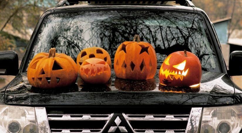 Four carved pumpkins are shown sitting atop the hood of a black Mitsubishi car.
