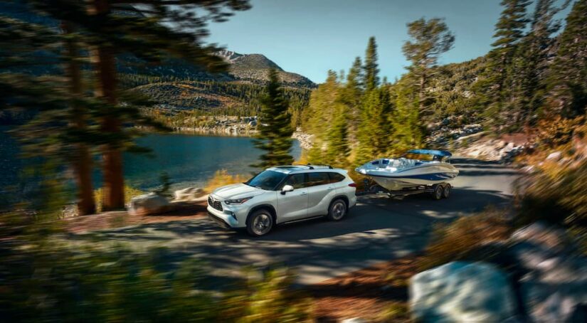 A white 2023 Toyota Highlander XLE is shown towing a boat through a forest after visiting a Toyota dealer in the city.