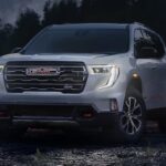 A silver 2024 GMC Acadia. is shown parked off-road with the headlights on.