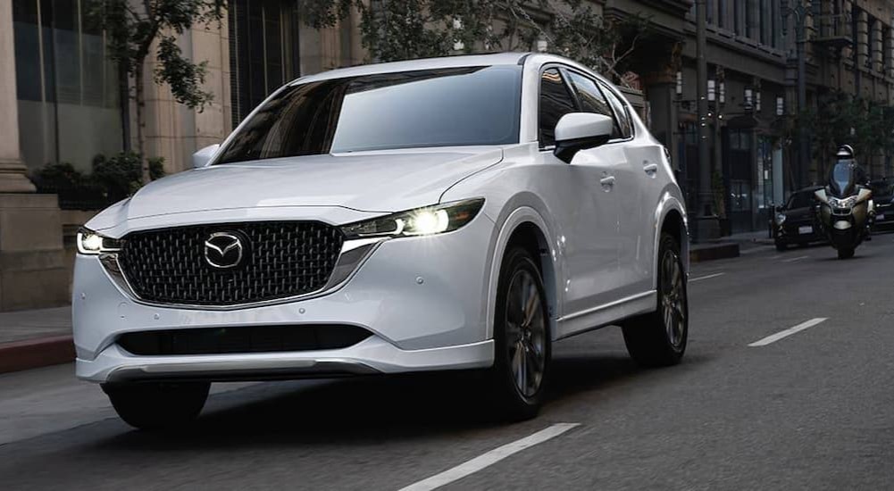 A white 2024 Mazda CX-5 is shown driving on a city street.