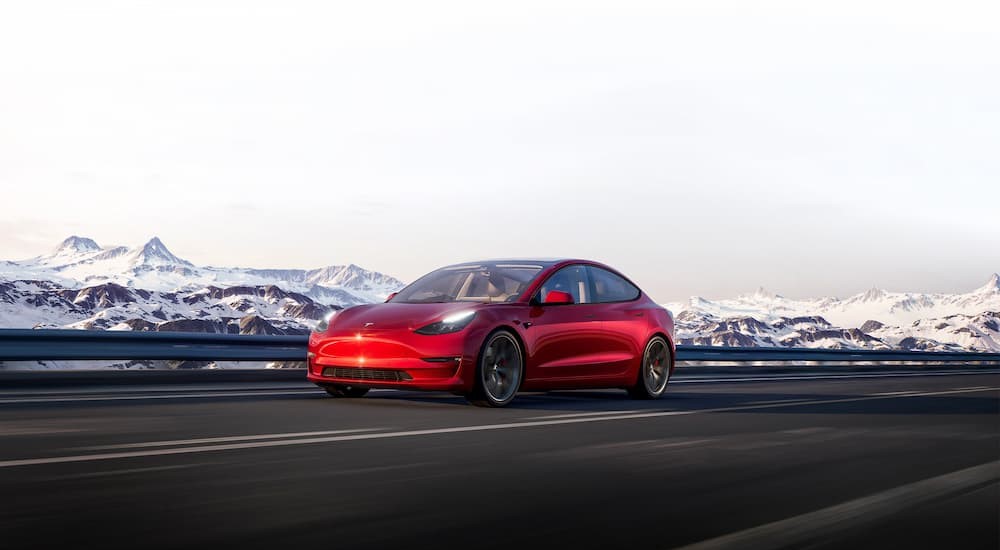 A red 2024 Tesla Model 3 Performance is shown driving on a highway near snowy mountains.