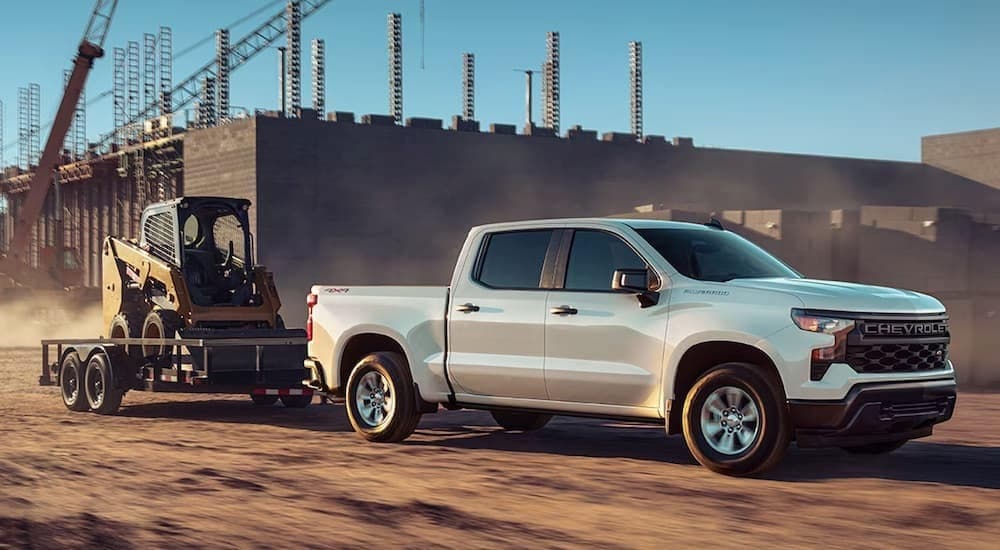 A white 2024 Chevy Silverado 1500 is shown towing a trailer on a construction site.