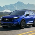 A blue 2024 Honda CR-V Sport Touring Hybrid is shown driving on a highway near mountains.