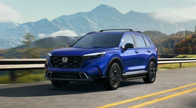 A blue 2024 Honda CR-V Sport Touring Hybrid is shown driving on a highway near mountains.
