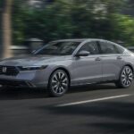 A grey 2023 Honda Accord Touring Hybrid is shown driving in a city after visiting a used car dealership.