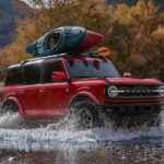 A red 2024 Ford Bronco Outer Banks is shown driving over a shallow river after visiting a car dealership.
