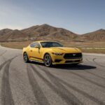 A yellow 2024 Ford Mustang parked on an empty road in front of mountains.