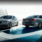 Two popular used cars for sale, two silver 2020 BMW 5 Series, are shown parked near a structure.
