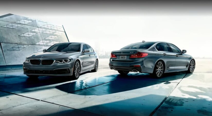 Two popular used cars for sale, two silver 2020 BMW 5 Series, are shown parked near a structure.