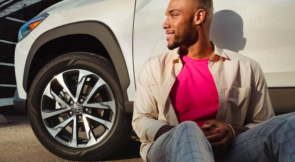 A man is shown sitting near the front wheel of a parked white 2022 Toyota Corolla Cross XLE.
