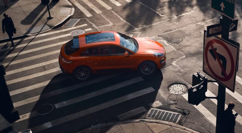 An orange 2024 Porsche Macan GTS is shown driving on an intersection.
