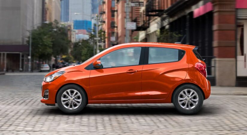 An orange 2021 Chevy Spark is shown driving on a city street after visiting a used Chevy dealer.