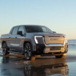 A silver 2024 GMC Sierra EV Denali Edition 1 driving on a shoreline after viewing trucks for sale.