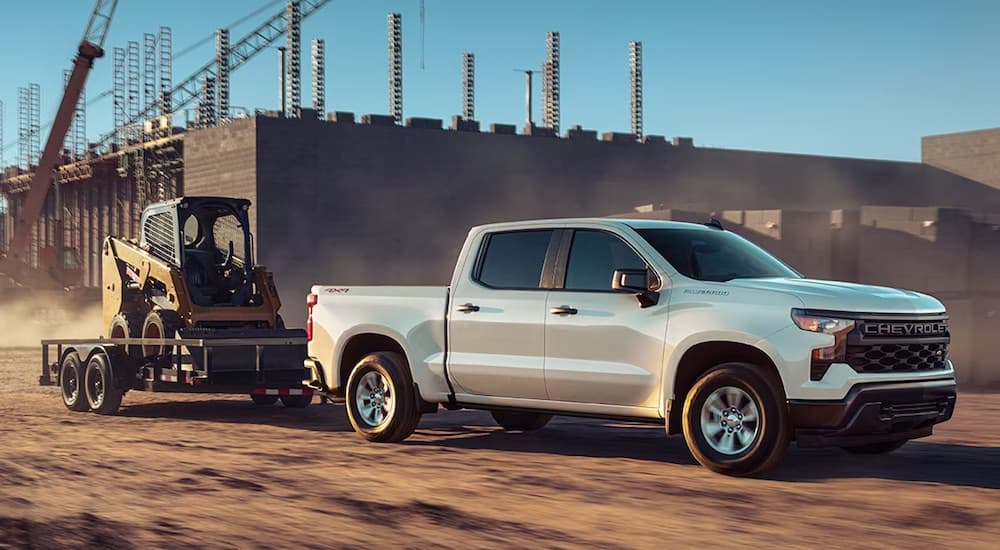 A white 2023 Chevy Silverado 1500 is shown from the side while towing a skid-steer.