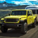 A popular Jeep Gladiator for sale, a yellow 2024 Jeep Gladiator Rubicon, is shown towing a trailer near a mountain.