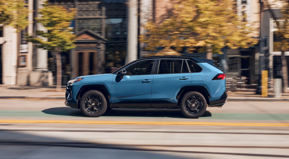 Side view of a blue 2024 Toyota RAV4 Hybrid driving through a city.