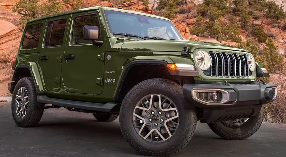 A green 2024 Jeep Wrangler Sahara parked in front of a rocky cliff.