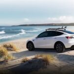 A white 2024 Tesla Model Y is shown parked on a beach near an ocean.