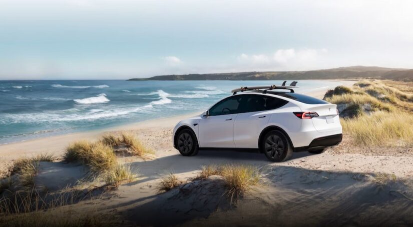 A white 2024 Tesla Model Y is shown parked on a beach near an ocean.