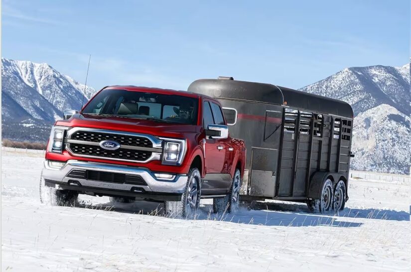 One of the more common used trucks for sale, a red 2023 Ford F-150 Powerboost Hybrid, is shown towing a trailer through a snowy field.