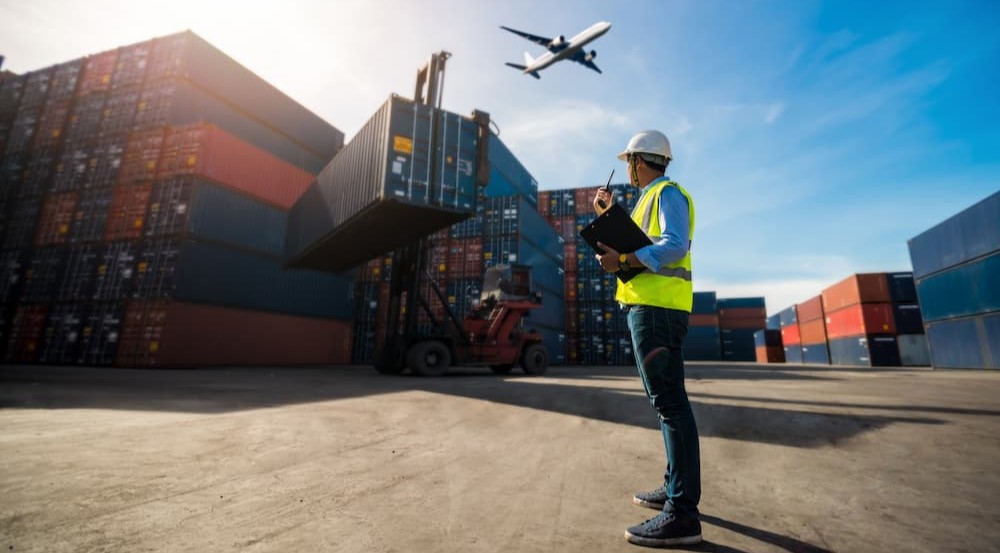 A person is shown standing near many shipping containers.