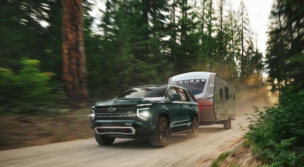 A green 2025 Chevy Tahoe Z71 towing a camper on a dirt road in the woods.