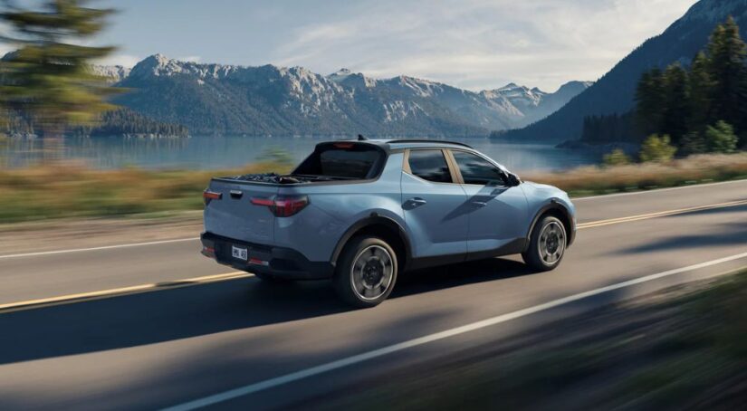 Rear view of a blue 2025 Hyundai Santa Cruz Limited driving past mountains and a lake.