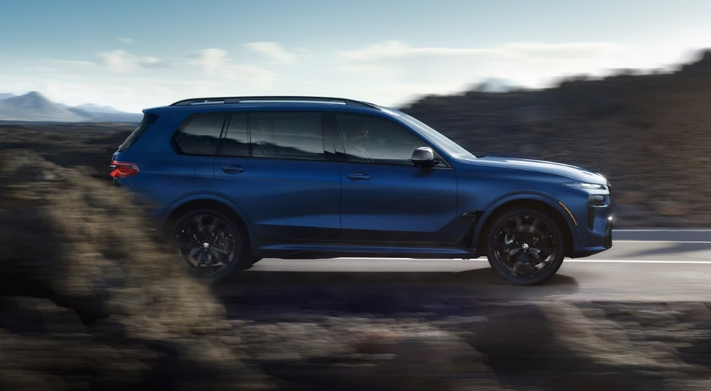 A blue 2026 BMW X7 M60i is driving past rocks.