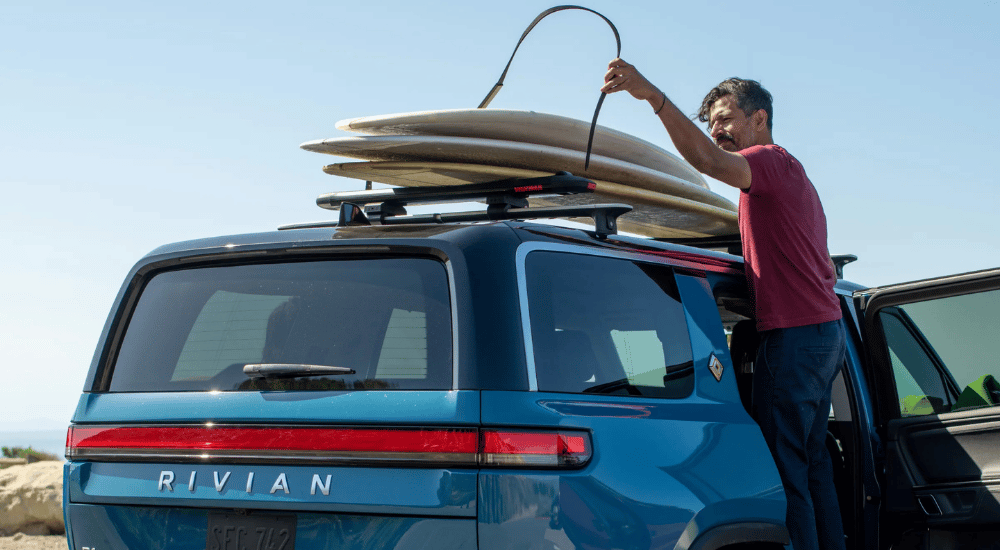 A person strapping surfboards on top of a blue 2026 Rivian R1S